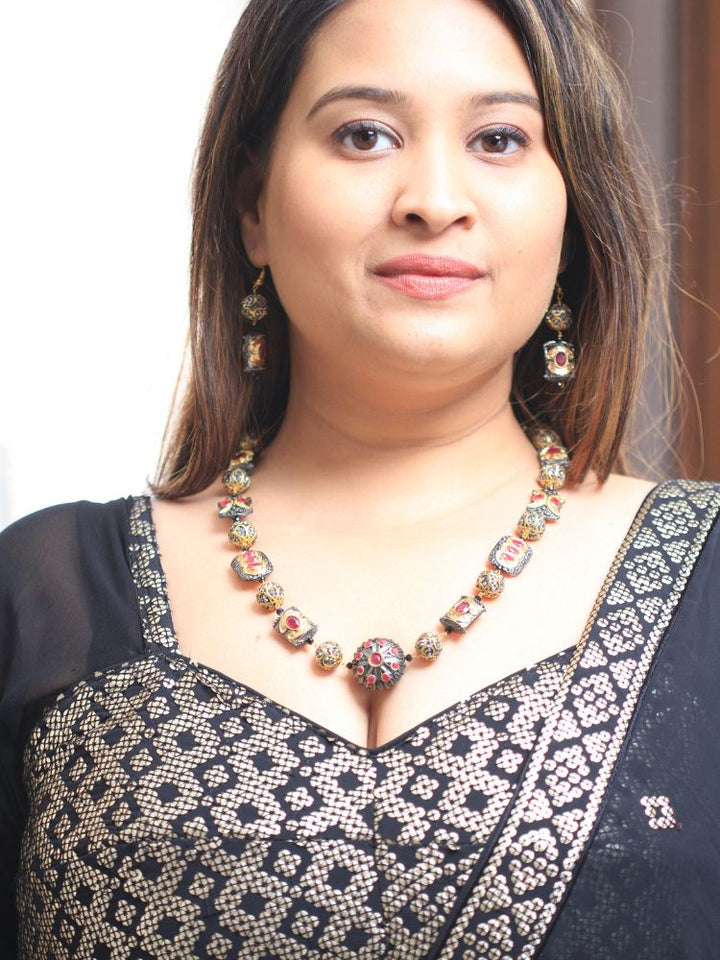 The Black Trisha Necklace Set is a decorative statement accessory with gold and black beads, square accents, and ruby and gold foil work, beautifully displayed on a girl in black attire with gold design on it. 