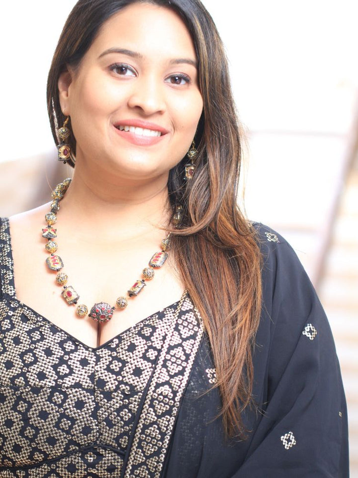 The Black Trisha Necklace Set is a decorative statement accessory with gold and black beads, square accents, and ruby and gold foil work, beautifully displayed on a girl in black attire with gold design on it. 