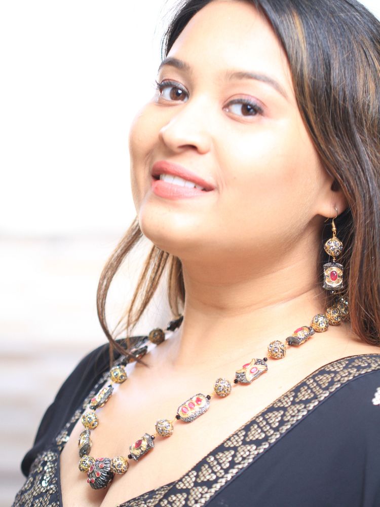 The Black Trisha Necklace Set is a decorative statement accessory with gold and black beads, square accents, and ruby and gold foil work, beautifully displayed on a girl in black attire with gold design on it. 