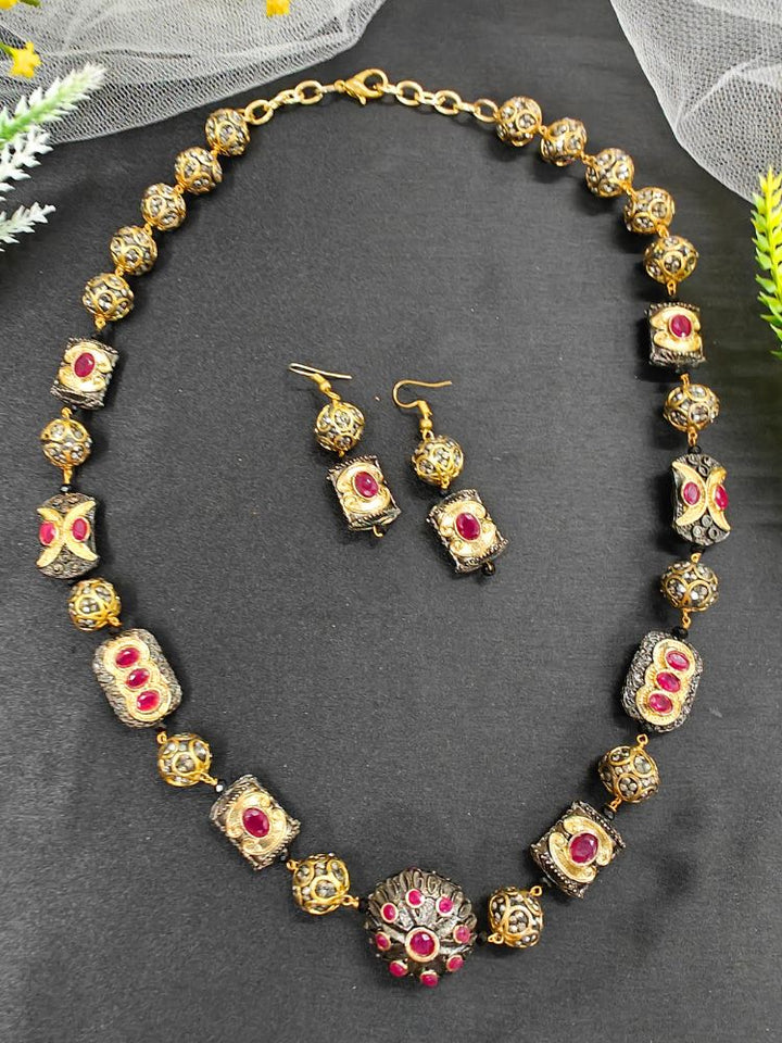 The Black Trisha Necklace Set is a decorative statement accessory with gold and black beads, square accents, and ruby and gold foil work, beautifully displayed on a black background. 