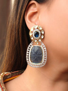 A close-up of a womans ear wearing Blue Fiona Earrings featuring a Blue carved stone, ornate American Diamonds and Kundan. Her dark hair is partially visible, and she wears a colorful embroidered garment.