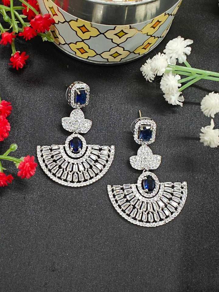 The Navy Blue Nora Earrings feature ornate fan-shaped designs with American Diamonds, displayed on a black surface near red and white flowers and a decorative bowl with floating elements.