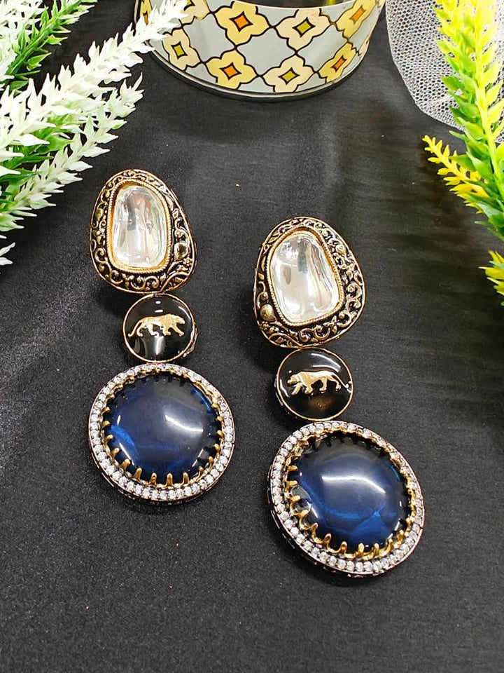 Blue Sabyasachi Inspired Earrings 6: Ornate blue earrings with gold and silver-toned metal, colored stones, round blue centers accented by American Diamonds, and a gold lion design at the center, presented on a black surface with decorative plants. Sabyasachi Earrings. American Diamond Earrings. Long Earrings. Party Earrings. 