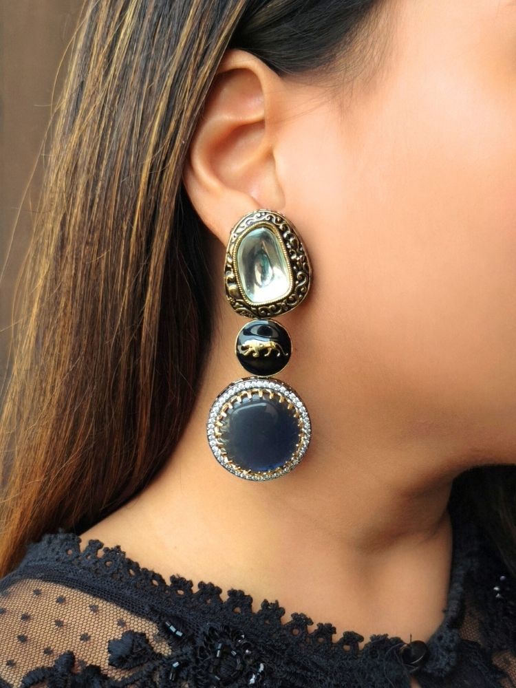 A close-up of a womans ear wearing Blue Sabyasachi Inspired Earrings 6, featuring a gold oval with a reflective stone, a black round section, and a circular pendant adorned with American Diamonds around a light brown center. Sabyasachi Earrings. American Diamond Earrings. Long Earrings. Party Earrings. 