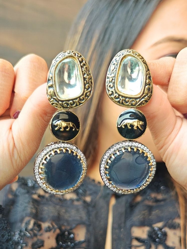 A woman in a black lace top holds up the Sabyasachi Inspired Earrings 6—Ornate blue earrings with gold and silver-toned metal, colored stones, round blue centers accented by American Diamonds, and a gold lion design at the center making them the perfect statement piece for a cocktail gown. Sabyasachi Earrings. American Diamond Earrings. Long Earrings. Party Earrings. 