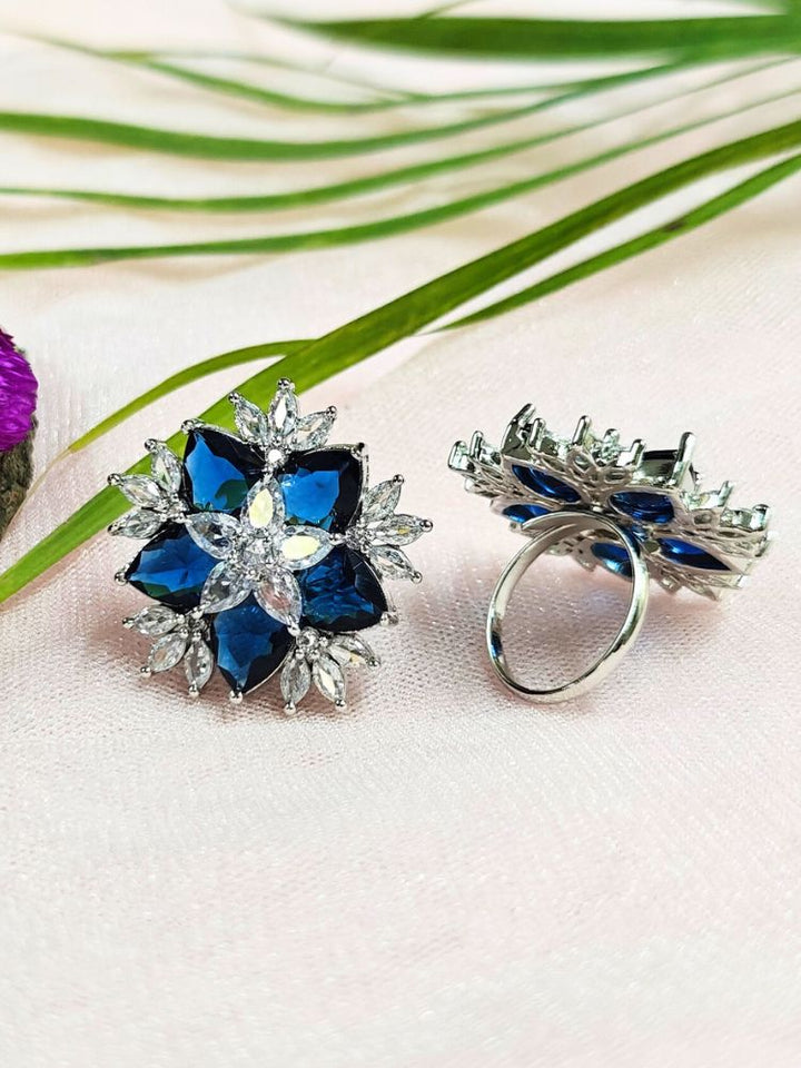 Two ornate, adjustable Blue Snowflake Rings with large Blue gemstone and american diamonds in floral designs—ideal as cocktail rings—are displayed on a light pink surface with green leaves and a purple flower nearby.