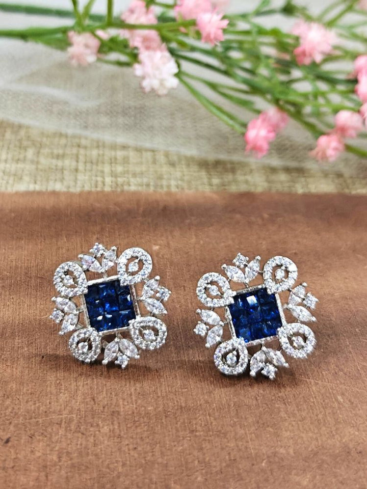 The Blue Zuri Earrings feature ornate navy blue square gemstones at the center, accented by intricate silver designs and sparkling American diamonds, displayed on a brown surface with pink flowers in the background.