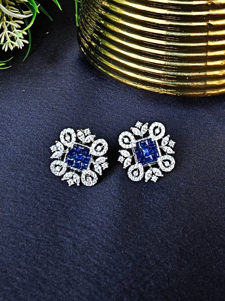 The Blue Zuri Earrings feature ornate navy blue square gemstones at the center, accented by intricate silver designs and sparkling American diamonds, displayed on a Black surface with White flowers in the background.