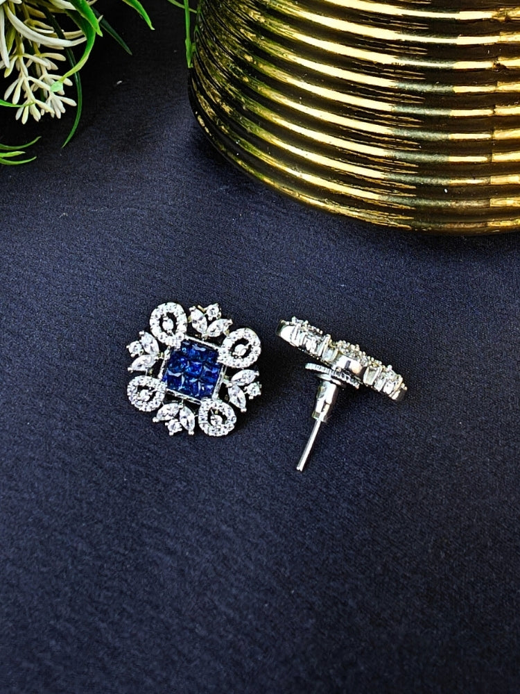 The Blue Zuri Earrings feature ornate navy blue square gemstones at the center, accented by intricate silver designs and sparkling American diamonds, displayed on a Black surface with White flowers in the background.