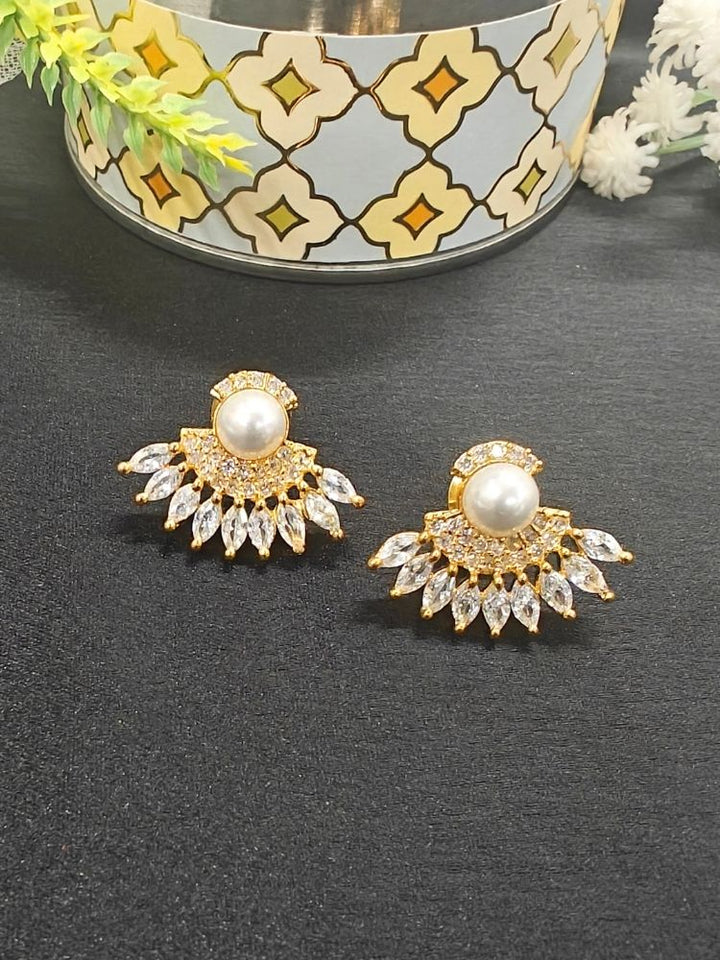 The Brooke Earrings are gold and white, featuring large pearl centers surrounded by small American Diamond stones in a fan shape, displayed on a black surface with a decorative container and flowers in the background.