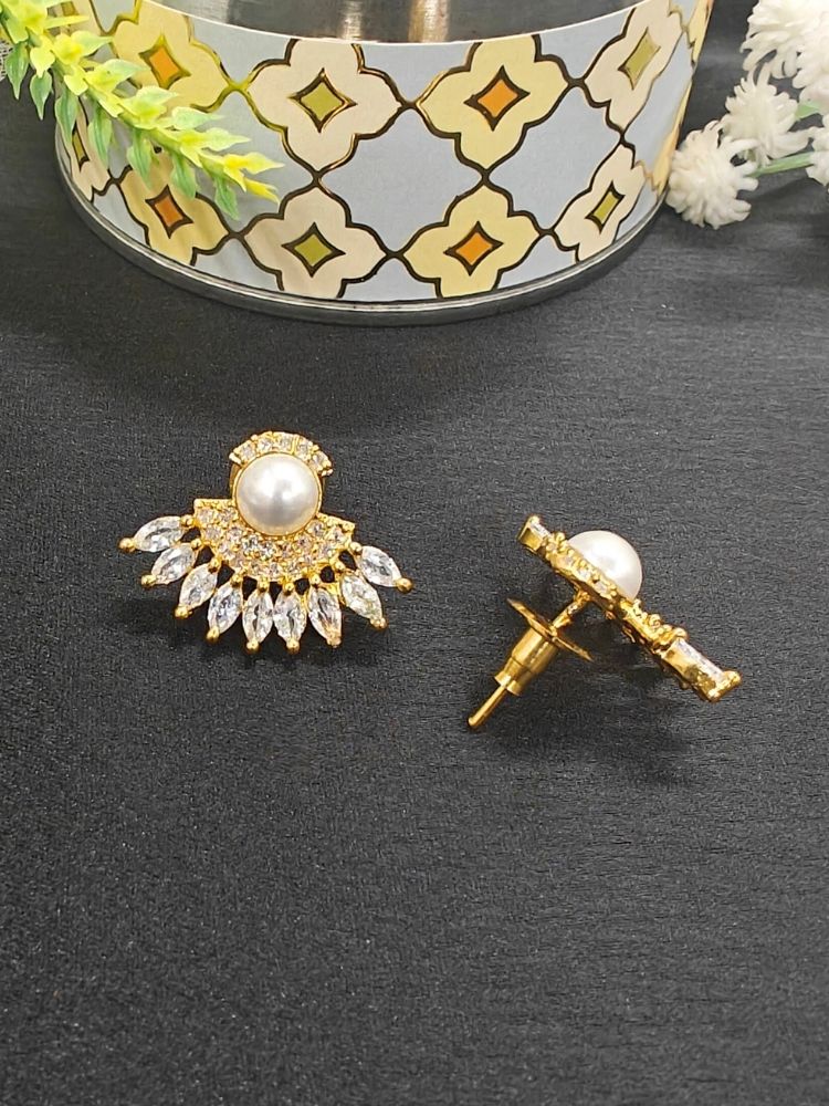 The Brooke Earrings are gold and white, featuring large pearl centers surrounded by small American Diamond stones in a fan shape, displayed on a black surface with a decorative container and flowers in the background.