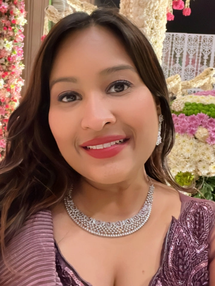 A woman wearing a shimmering purple cocktail gown and the Camille Lilac American Diamond Cocktail Necklace Set smiles for a selfie, with white decorative trees and vibrant floral arrangements in the background. 