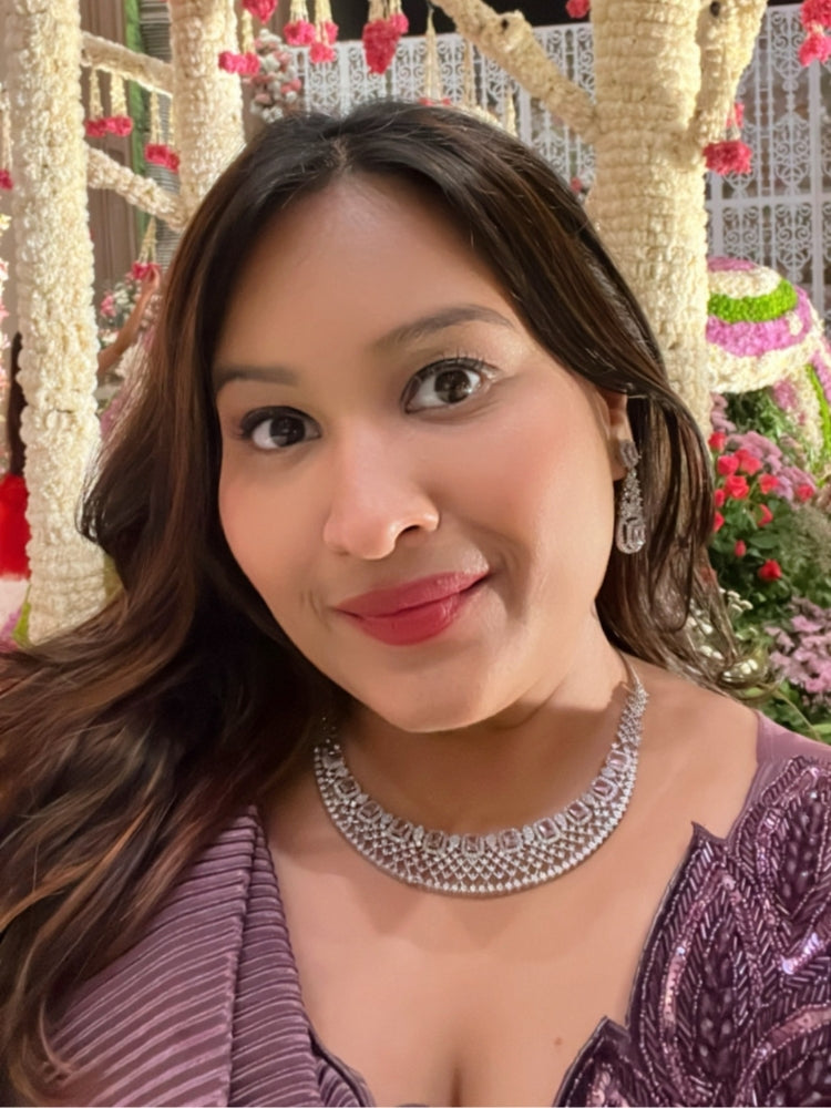 A woman wearing a shimmering purple cocktail gown and the Camille Lilac American Diamond Cocktail Necklace Set smiles for a selfie, with white decorative trees and vibrant floral arrangements in the background. 