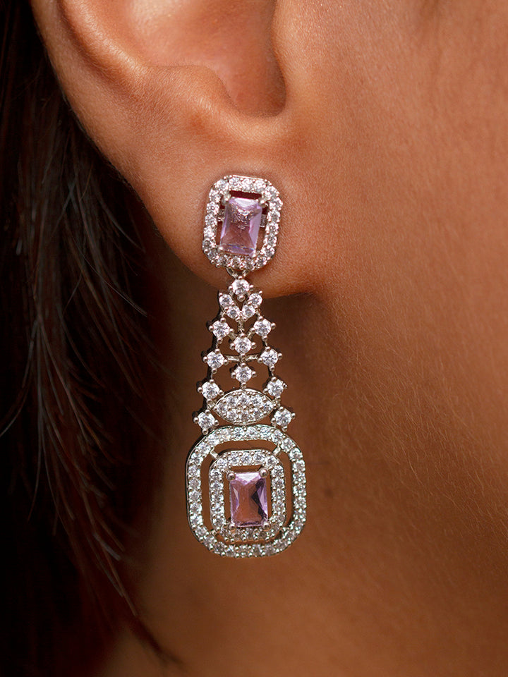 A woman wearing a shimmering purple cocktail gown and the Camille Lilac American Diamond Cocktail Necklace Set smiles for a selfie, with white decorative trees and vibrant floral arrangements in the background. 
