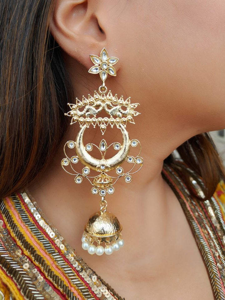 A close-up of a womans ear adorned with Carol Earrings, showcasing Kundan work, pearls, American Diamonds, and floral-circular designs with a Jhumka at the bottom. Giving a perfect mix of chandbali and Jhumka look. She wears a colorful embroidered outfit inspired by traditional Indian attire.
