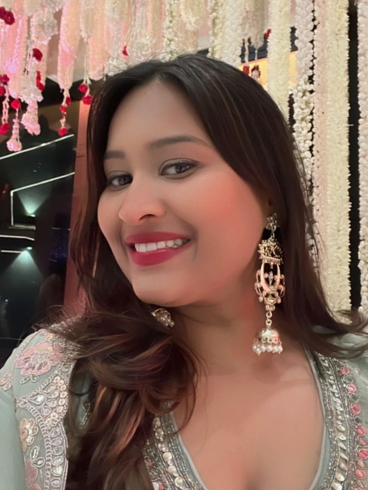 A close-up of a womans ear adorned with Carol Earrings, showcasing Kundan work, pearls, American Diamonds, and floral-circular designs with a Jhumka at the bottom. Giving a perfect mix of chandbali and Jhumka look. She wears a colorful embroidered outfit inspired by traditional Indian attire.