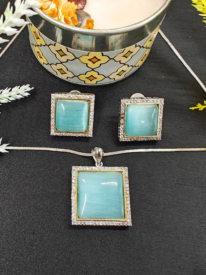 The Carter Blue Pendant Set features a square blue pendant and matching earrings with silver frames surrounded by American diamonds, displayed on black near a candle and flowers—an elegant ethnic jewelry piece for any occasion. Perfect for casual weal. Casual pendant set. 