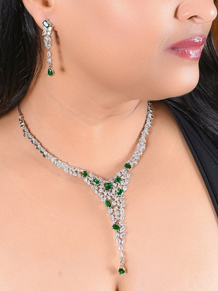 A woman wears the Celestia Green Necklace Set—an American Diamond necklace and matching green gemstone earrings—with a black top. The elegant set is perfect for weddings, complemented by her glossy pink lipstick.