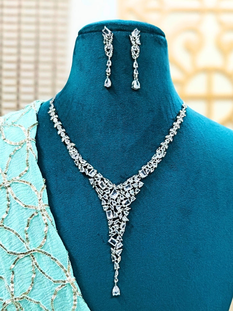 A mannequin showcases the Celestia White Necklace Set—featuring sparkling American diamonds in a 'V' shape necklace, and matching dangling earrings—paired with a light blue and gold embroidered saree draped over one shoulder.