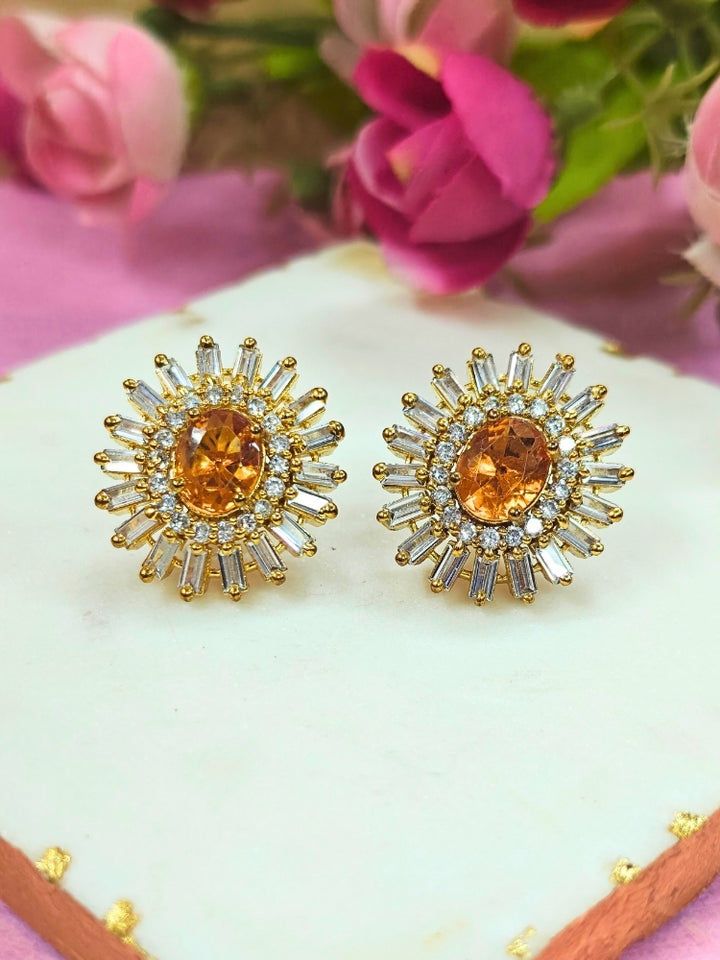 The Celosia Earrings feature an oval orange gemstone at the center, accented by a sunburst of clear stones and American Diamonds, set in gold and elegantly displayed on a white surface with pink flowers in the background. American Diamond Studs. Studs. Stud earrings. Everyday use earrings. 