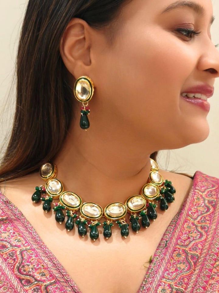 A woman in a pink patterned outfit smiles and looks to the side, wearing the elegant Chaitali Necklace Set—featuring green tumble stones hanging with big oval shaped Kundans along with matching earrings—ideal for any wedding event.