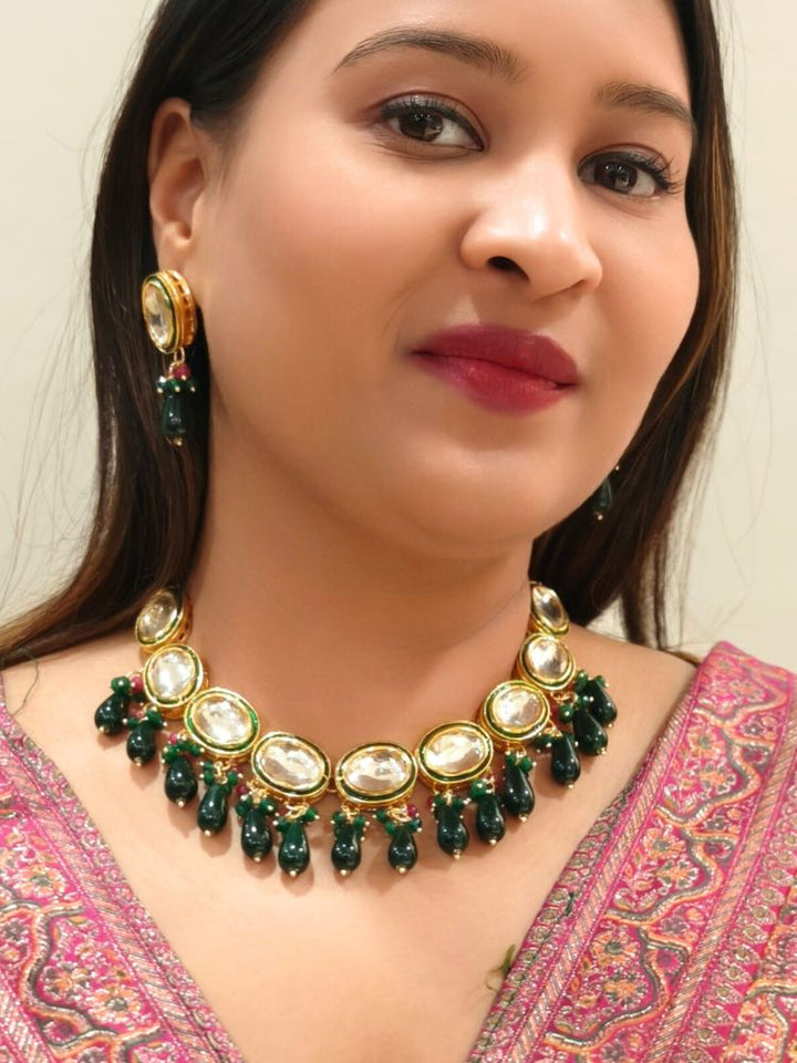 A woman in a pink patterned outfit smiles and looks to the side, wearing the elegant Chaitali Necklace Set—featuring green tumble stones hanging with big oval shaped Kundans along with matching earrings—ideal for any wedding event.