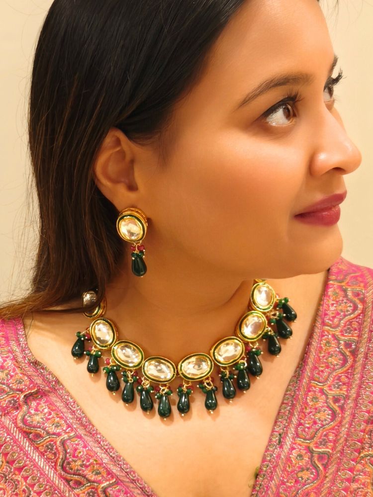 A woman in a pink patterned outfit smiles and looks to the side, wearing the elegant Chaitali Necklace Set—featuring green tumble stones hanging with big oval shaped Kundans along with matching earrings—ideal for any wedding event.