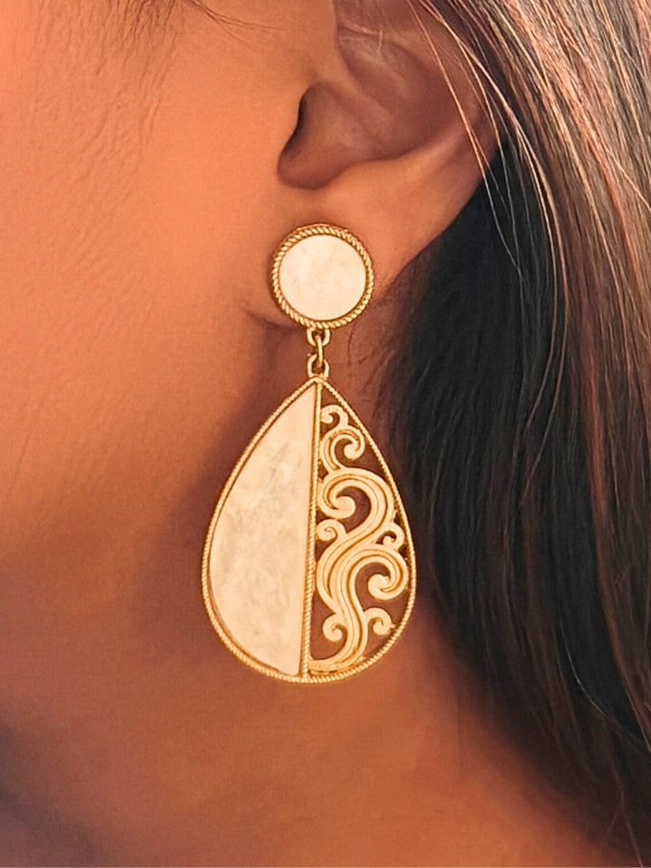 A close-up of an ear wearing the Chandsona Earrings—large teardrop earrings in Mother of Pearl and gold, with a round stud and ornate swirling detail. These modern, elegant White & Gold Earrings create a striking look.