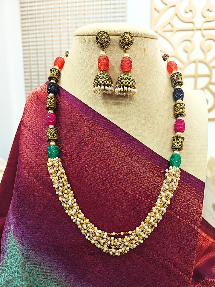 A mannequin showcases the Charvi Necklace Set—an elegant gold piece with oxidized brass beads, multicolored stones, with multiple strands of pearls, paired with matching jhumka earrings and styled with a maroon and purple saree—perfect for adding flair to any wedding ceremony.