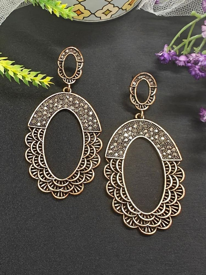 The Christine Earrings are ornate, oval-shaped brass oxidized long earrings with intricate patterns and small crystal stones, displayed on a black surface with green and purple flowers nearby.