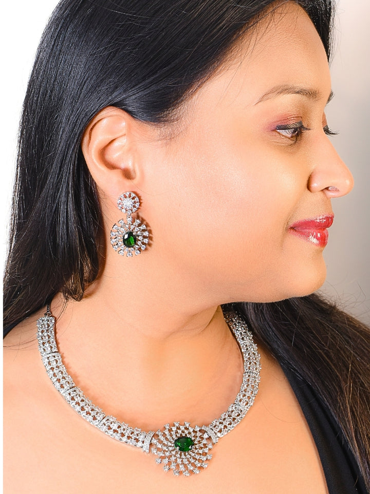 A woman adorns herself with the Cressida Green Necklace Set, featuring sparkling American Diamond necklace,  highlighted by a striking green gemstone in the center. This necklace set comes along with matching earrings. The jewelry intricate design elegantly enhances her look.