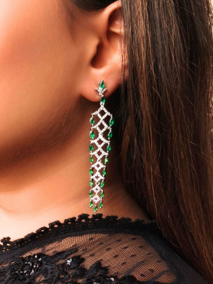 A close-up of a womans ear wearing Cyra Earrings—long, elegant green colored stone earrings with American diamonds and a silver lattice design. She has straight brown hair and a black lace top, making them ideal for your favorite party dress.