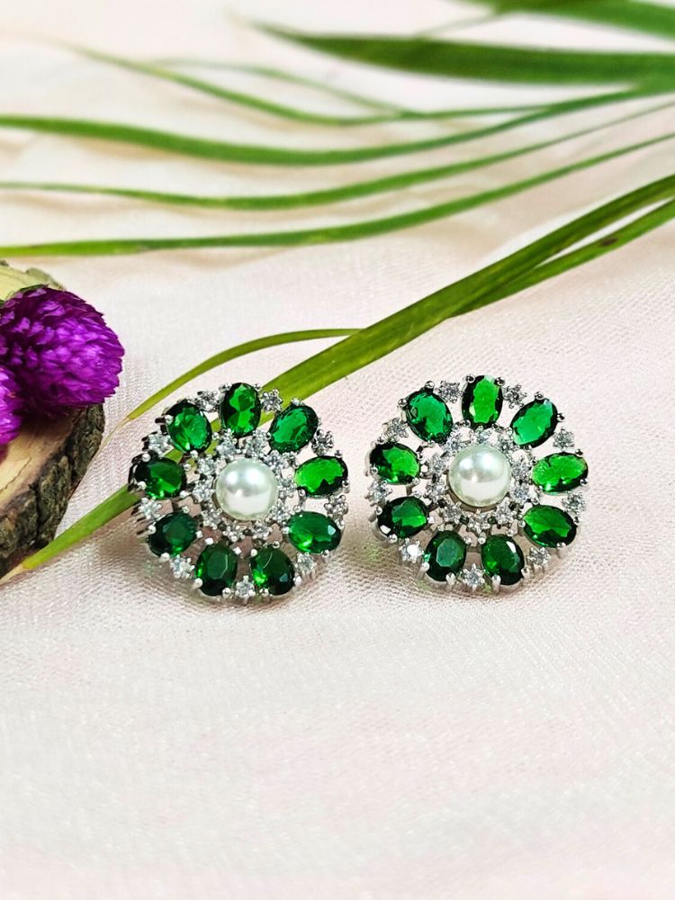 The Dark Green Blossom Earrings showcase a central white pearl encircled by alternating Dark Green stones and American diamonds, elegantly displayed on soft pink fabric with green leaves and a purple flower.
