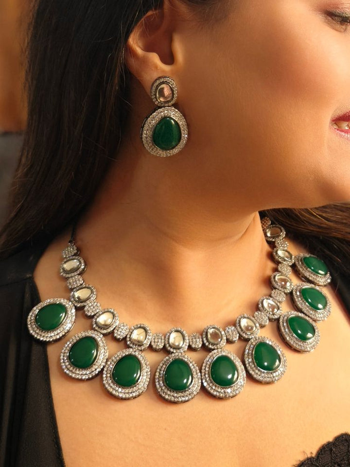 A woman with brown hair down smiles to the side, wearing the Dark Green Elizabeth Necklace Set featuring a statement Victorian-style necklace and matching earrings with large colorful stones, paired with a sparkly dark top.