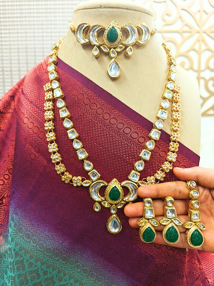 The Dark Green Nadira Necklace Set, features a tripple layered necklace in which there is a seperate choker made with carved stone in the center, along with kunda and pearl detailing on the side. The second necklace is a 2-layered necklace made with with curved stone in the center, along with kundan detailing all along the necklace. It comes with matching earrings. It is displayed on a mannequin in a maroon and teal saree; a hand holds the earrings.