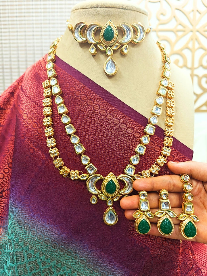 The Dark Green Nadira Necklace Set, features a tripple layered necklace in which there is a seperate choker made with carved stone in the center, along with kunda and pearl detailing on the side. The second necklace is a 2-layered necklace made with with curved stone in the center, along with kundan detailing all along the necklace. It comes with matching earrings. It is displayed on a mannequin in a maroon and teal saree; a hand holds the earrings.