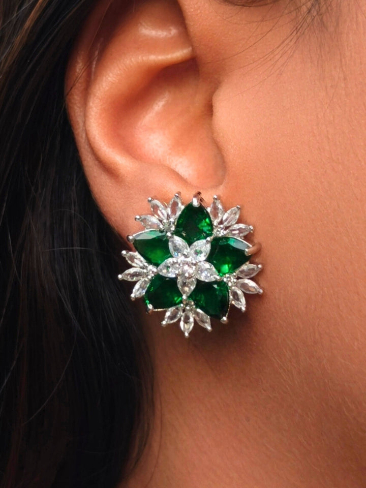 A close-up of an ear wearing Dark Green Snowflake stud Earrings, featuring flower-shaped earring with dark green gemstone petals and american diamonds outlining each petal and the center. Snowflake shaped stud earrings. 