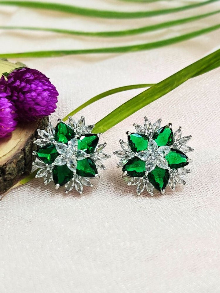 The Dark Green Snowflake Earrings showcase Dark Green colored stones along with American Diamonds in a starburst design, perched on a soft pink fabric with green leaves and a Purple flower.