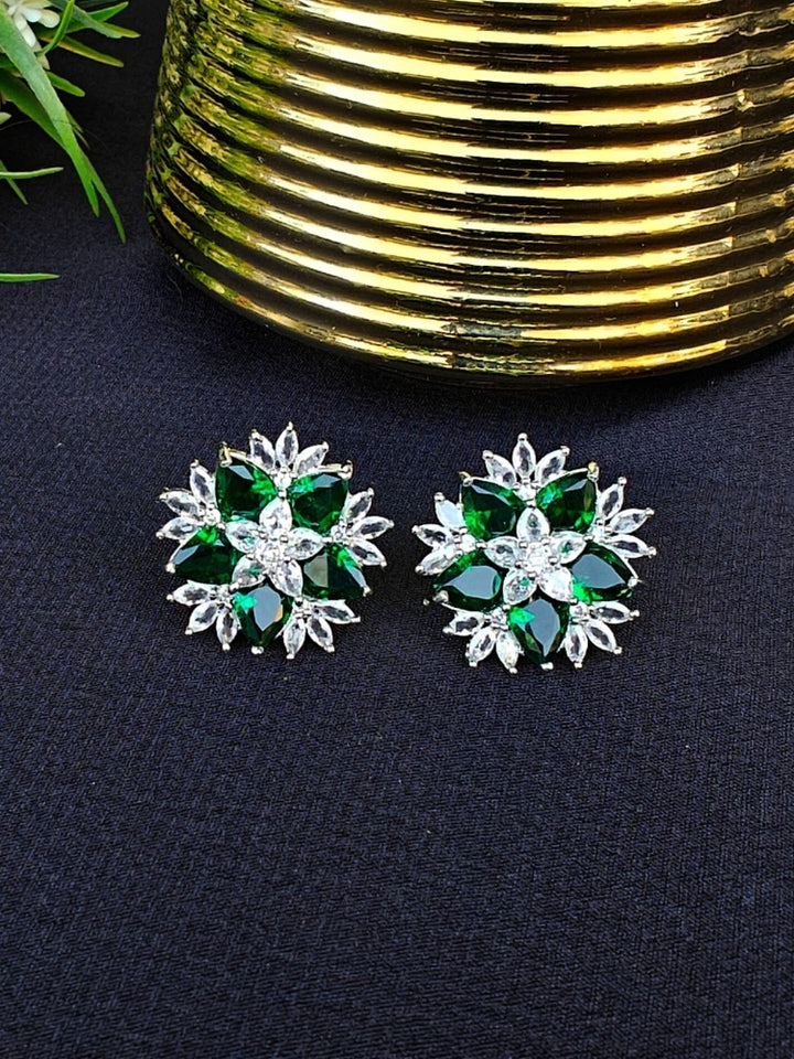 The Dark Green Snowflake Earrings showcase Dark Green colored stones along with American Diamonds in a starburst design, perched on a Black Surface with green leaves . 