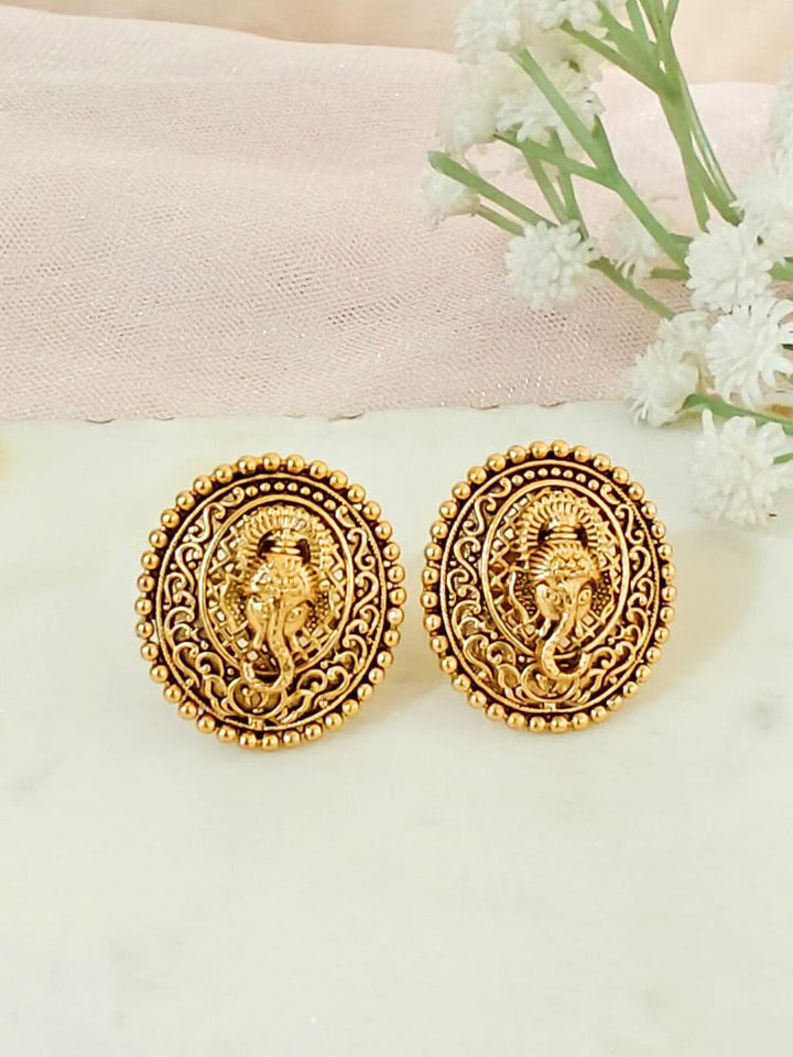The Deepali stud Earrings are round, traditional-style gold-toned brass earrings with intricate designs and a central Lord Ganesha motif, shown on a light surface with white flowers in the background.