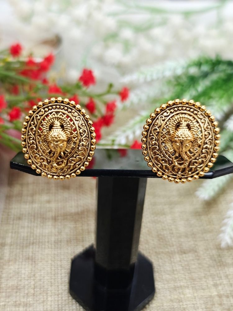 The Deepali stud Earrings are round, traditional-style gold-toned brass earrings with intricate designs and a central Lord Ganesha motif, shown on a light surface with white flowers in the background.