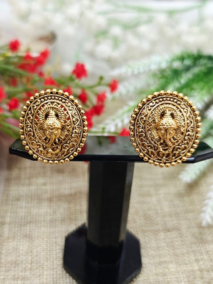 The Deepali stud Earrings are round, traditional-style gold-toned brass earrings with intricate designs and a central Lord Ganesha motif, shown on a light surface with white flowers in the background.