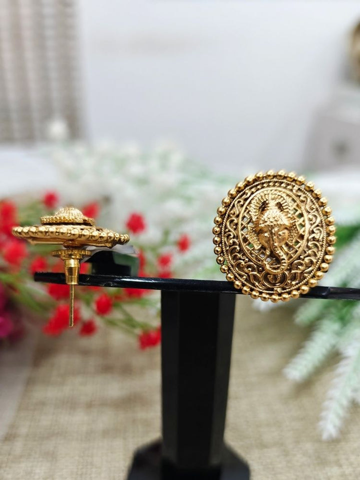 The Deepali stud Earrings are round, traditional-style gold-toned brass earrings with intricate designs and a central Lord Ganesha motif, shown on a light surface with white flowers in the background.