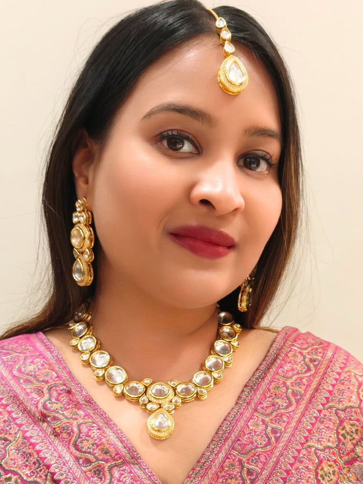 A woman wearing a Pink outfit and long brown hair smiles at the camera, showcasing the White Dhara Kundan Necklace Set—gold jewelry with White, and clear Monalisa stones—complete with necklace, earrings, and maang tikka.
