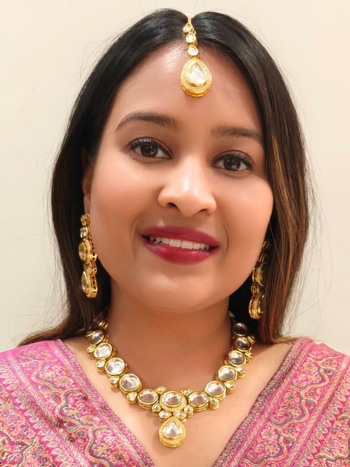 A woman wearing a Pink outfit and long brown hair smiles at the camera, showcasing the White Dhara Kundan Necklace Set—gold jewelry with White, and clear Monalisa stones—complete with necklace, earrings, and maang tikka.