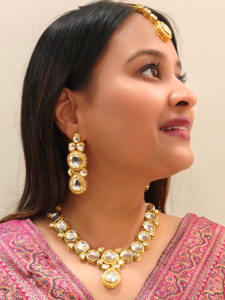 A woman wearing a Pink outfit and long brown hair smiles at the camera, showcasing the White Dhara Kundan Necklace Set—gold jewelry with White, and clear Monalisa stones—complete with necklace, earrings, and maang tikka.