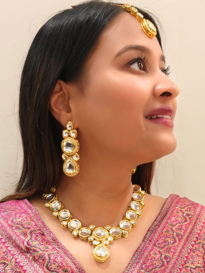 A woman wearing a Pink outfit and long brown hair smiles at the camera, showcasing the White Dhara Kundan Necklace Set—gold jewelry with White, and clear Monalisa stones—complete with necklace, earrings, and maang tikka.