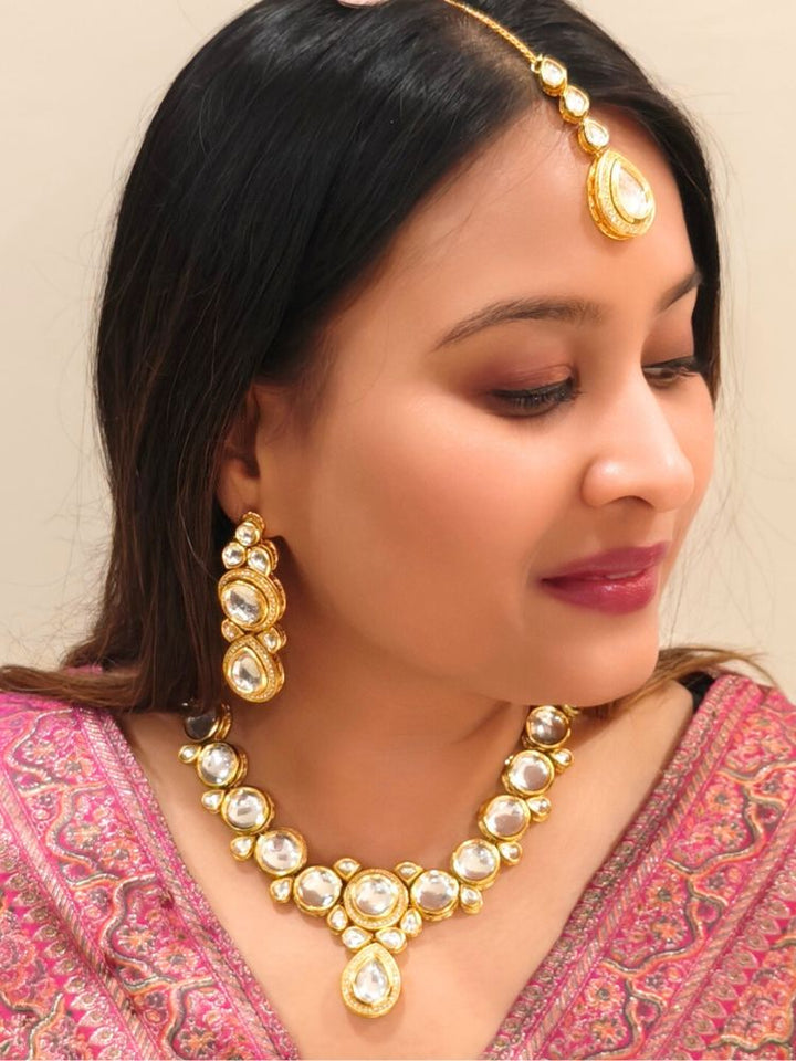 A woman wearing a Pink outfit and long brown hair smiles at the camera, showcasing the White Dhara Kundan Necklace Set—gold jewelry with White, and clear Monalisa stones—complete with necklace, earrings, and maang tikka.