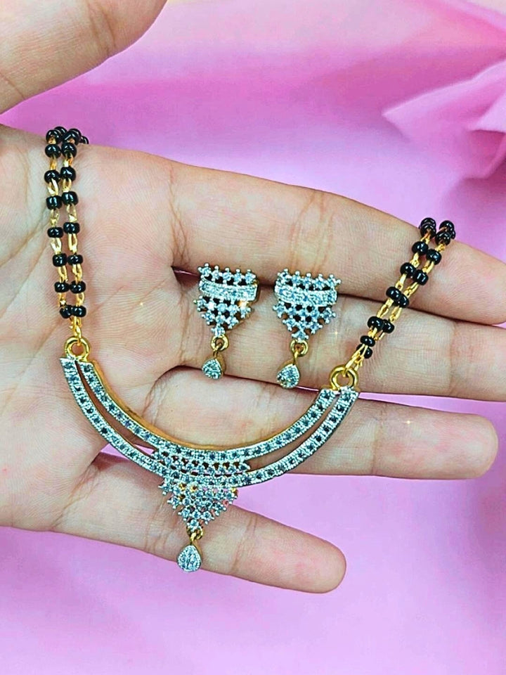 A hand holds the Diksha Mangalsutra—a gold necklace with black mangalsutra beads and matching earrings, pendant featuring sparkling American Diamonds—set against a soft pink background, perfect for your wedding ceremony dress. Diamond mangalsutra. Modern mangalsutra. 