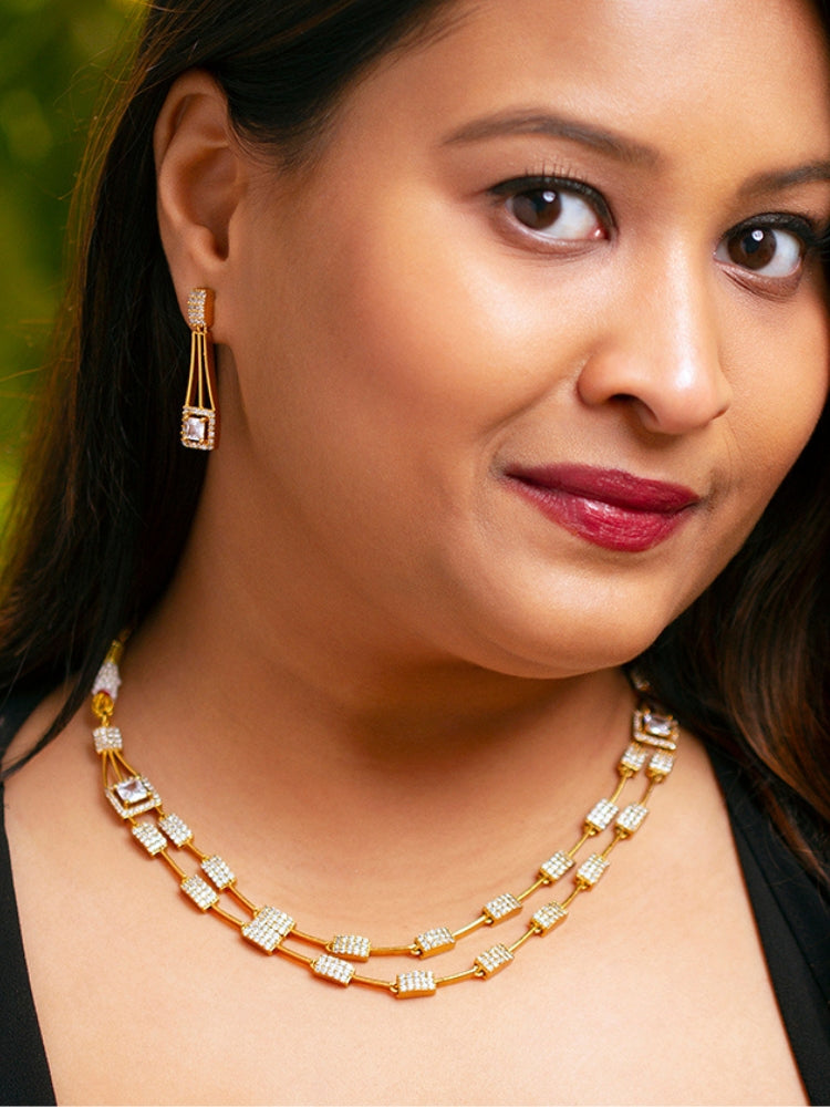 A woman wears the Diora White Necklace Set, a gold party jewelry set featuring a White 2 layer necklace set made with  American diamonds and White gemstones at both the corners, paired with matching earrings. Only her lower face and neck are visible.